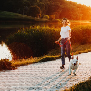 Happy female running with white dog on leash during walk at parkway with water and grass with trees on backlit background in evening